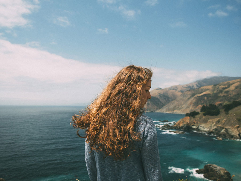 Garota de costas olhando para baixo, ao fundo uma paisagem com mar e rochas